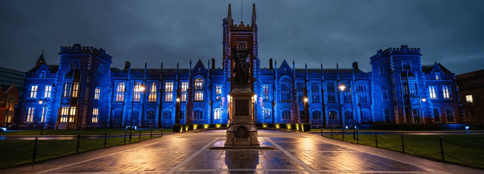 Front of Queen's University Lanyon building illuminated blue (The Lanyon Building features a grand, symmetrical red-brick façade with sandstone trim. It has a large arched entrance, tall narrow windows, and a prominent clock tower with a spire. Decorative stone carvings and battlements give it a castle-like appearance